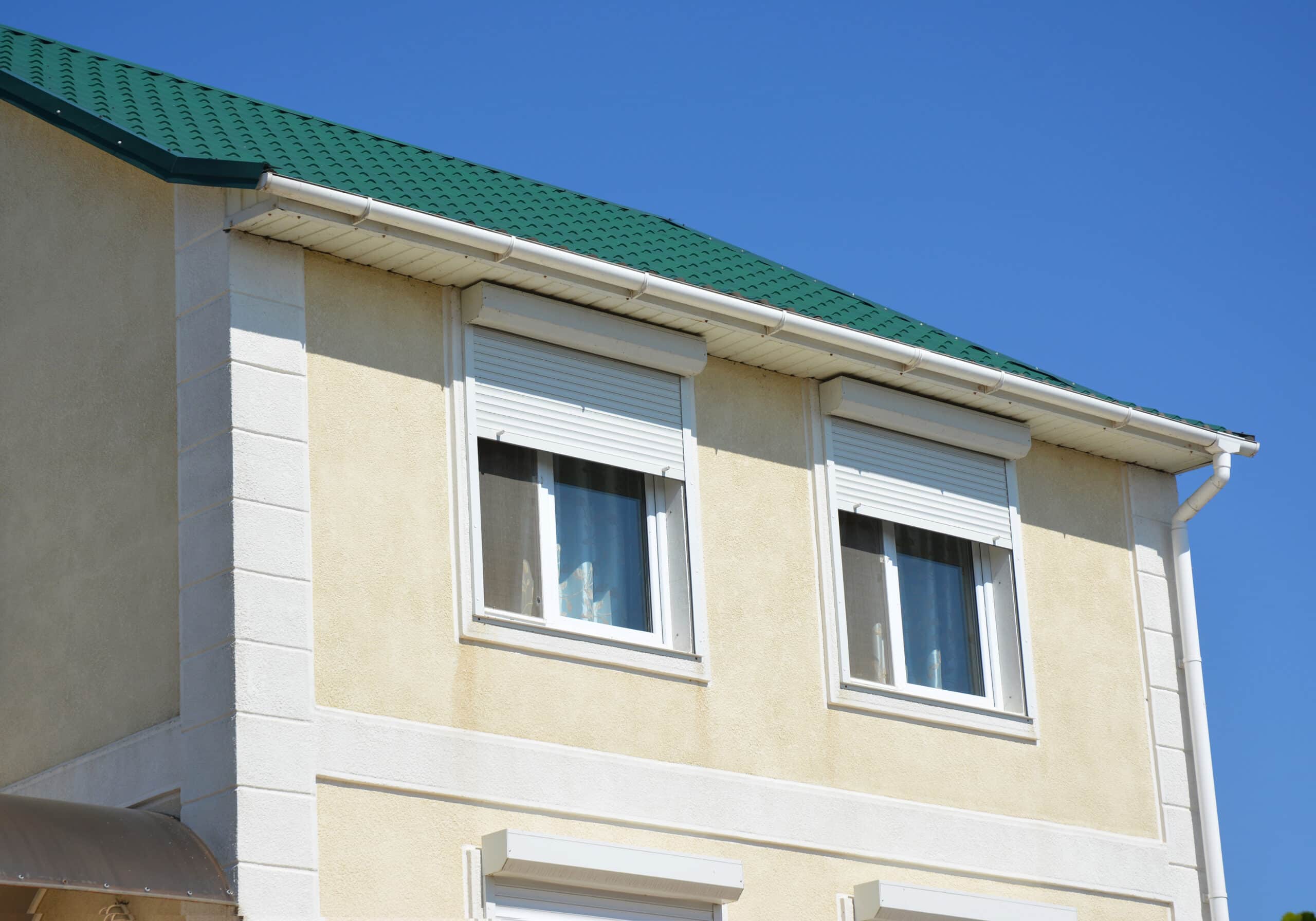 A close up of a stucco house with a metal green roof, rain gutters, soffit, fascia, and windows with white exterior pvc motorized window roller shutters.